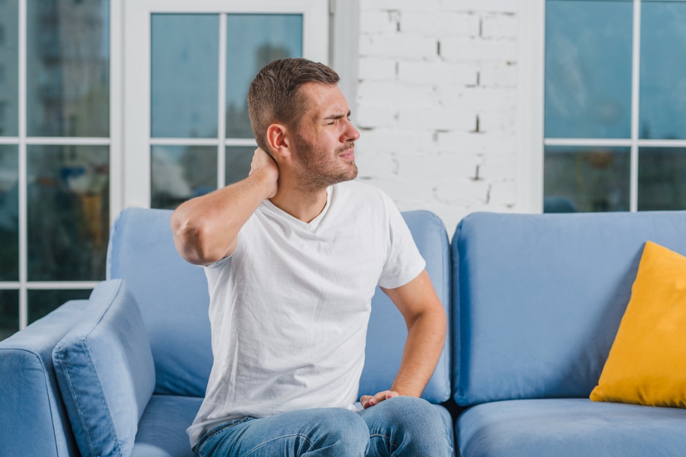 A man sits on a couch holding his neck in discomfort, representing everyday aches and effective Chronic Pain Management Delaware care solutions.